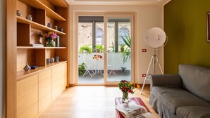 Una sala de estar moderna con un sofá gris, estanterías de madera con objetos decorativos, una lámpara de pie y vistas a un patio con plantas y una mesa a través de una puerta de cristal.