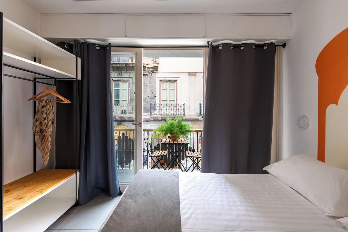 A bedroom with a bed, a wooden shelf, a potted plant, and a balcony with a view of buildings.