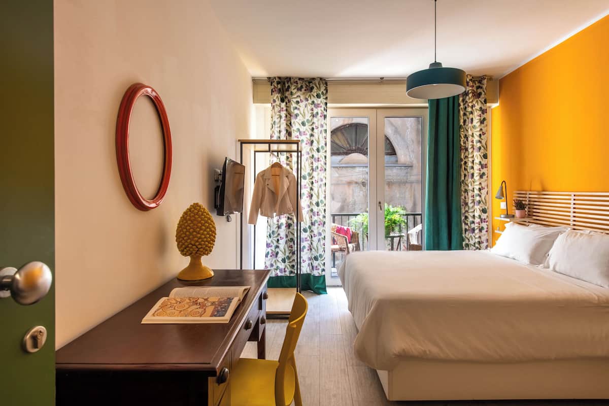 A hotel room with a bed, a desk with a chair, a coat rack, and a door.