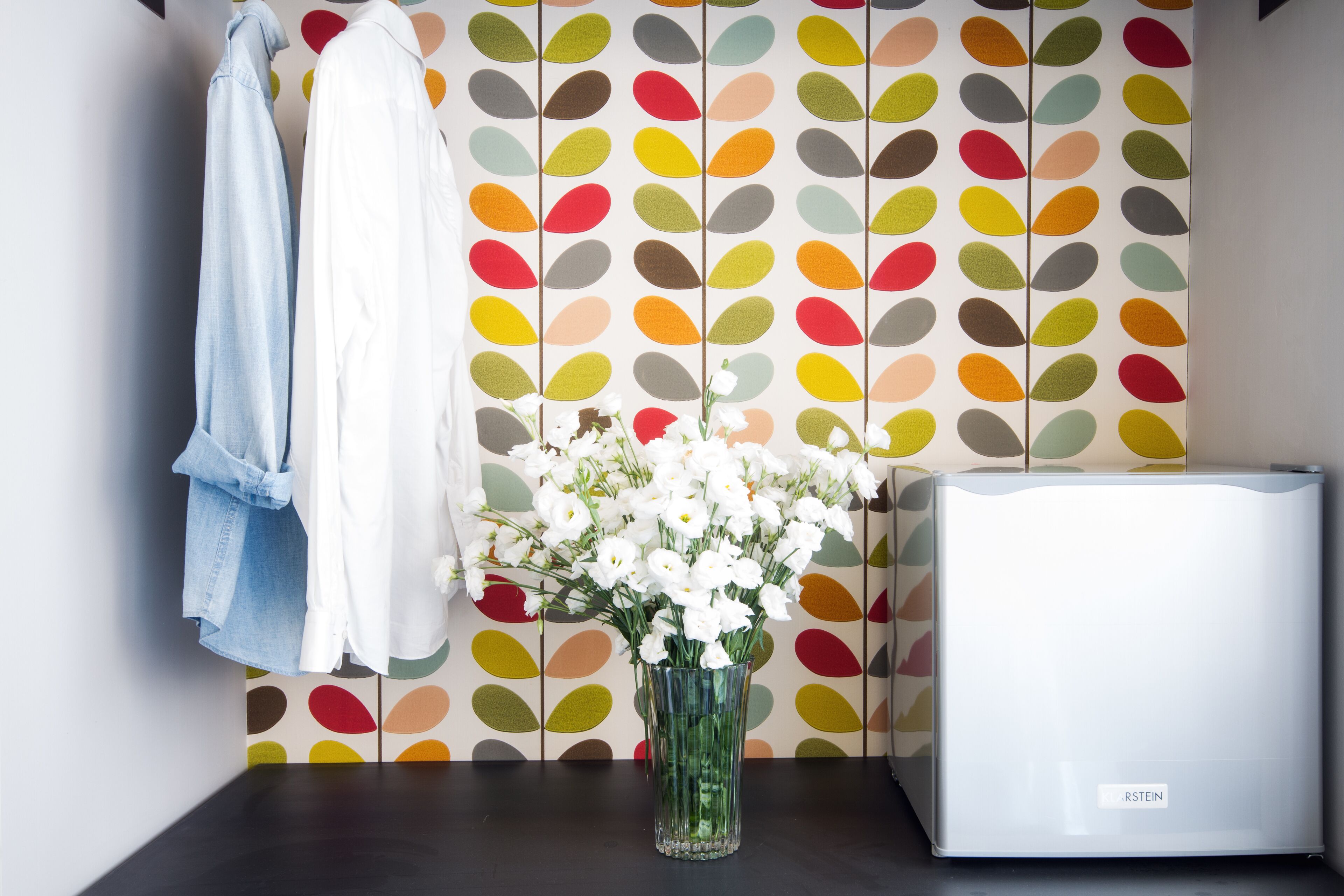 A room with a patterned wallpaper, a white appliance, and a vase with white flowers on a table.