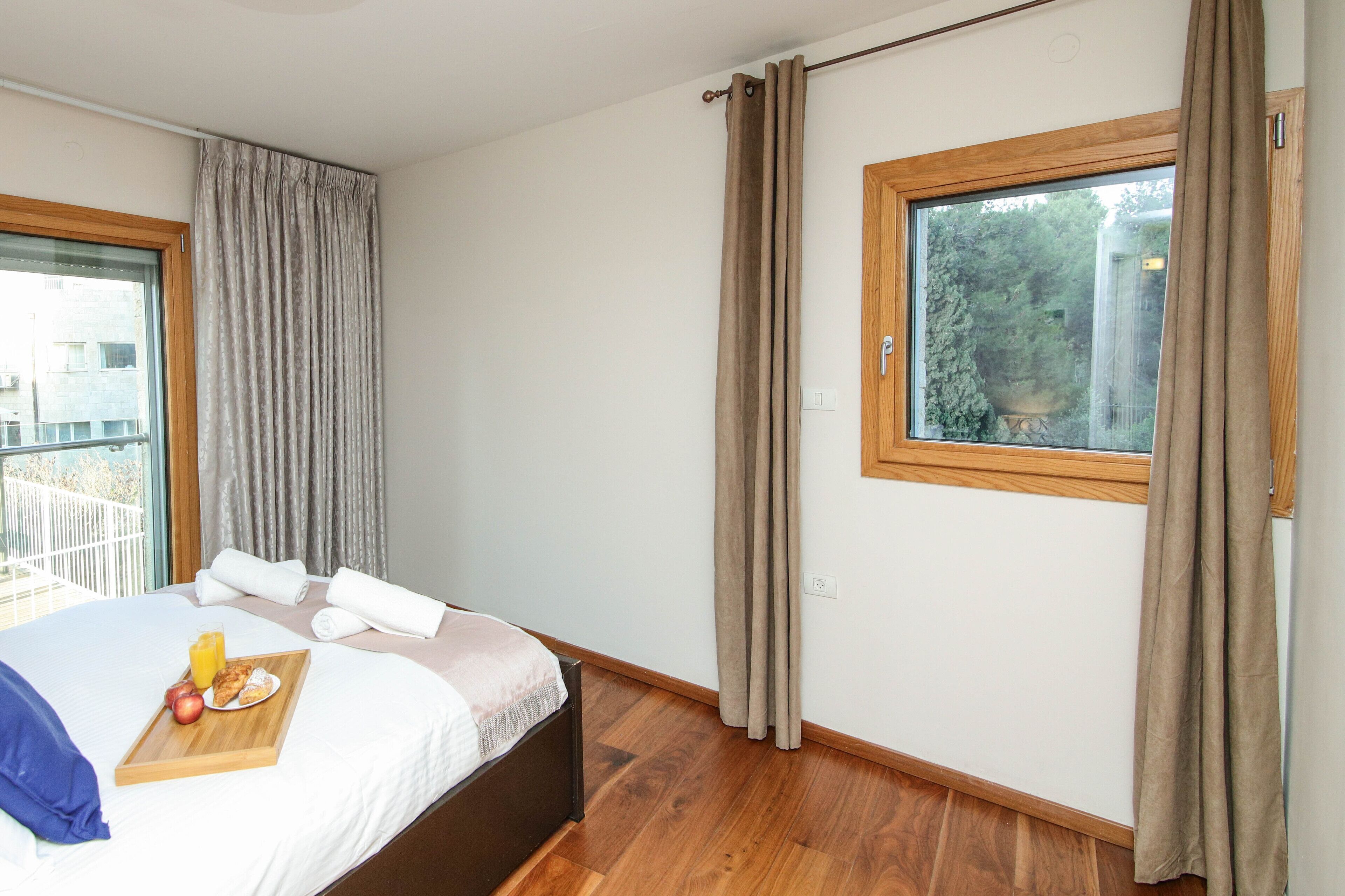 A bedroom with a bed, a wooden bedside table with a tray of breakfast, a window with curtains, and a view of greenery outside.