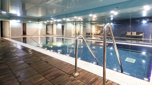 An indoor swimming pool with clear glass walls, wooden flooring, and metal railings.