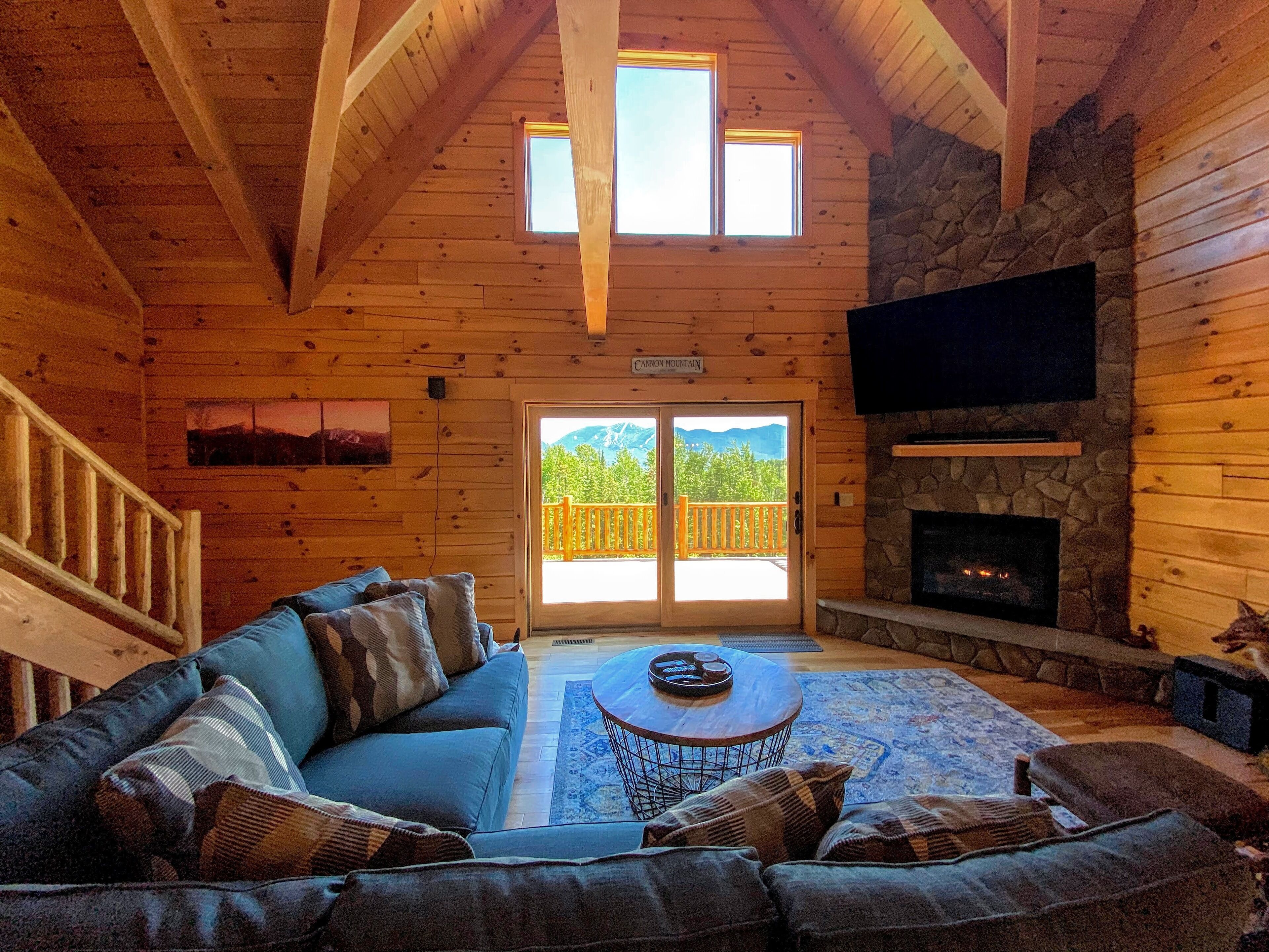L’intérieur chaleureux d’un chalet en bois, avec une cheminée en pierre, un grand canapé et un balcon offrant une vue imprenable sur les montagnes.