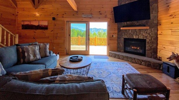 L’intérieur chaleureux d’un chalet avec une cheminée en pierre, un grand téléviseur fixé au mur, un plafond en bois et une vue sur les montagnes à travers les portes vitrées.