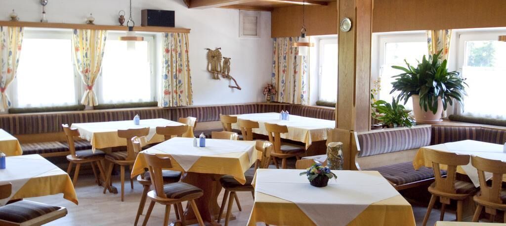 A restaurant with multiple tables and chairs, large windows with curtains, and a variety of plants.