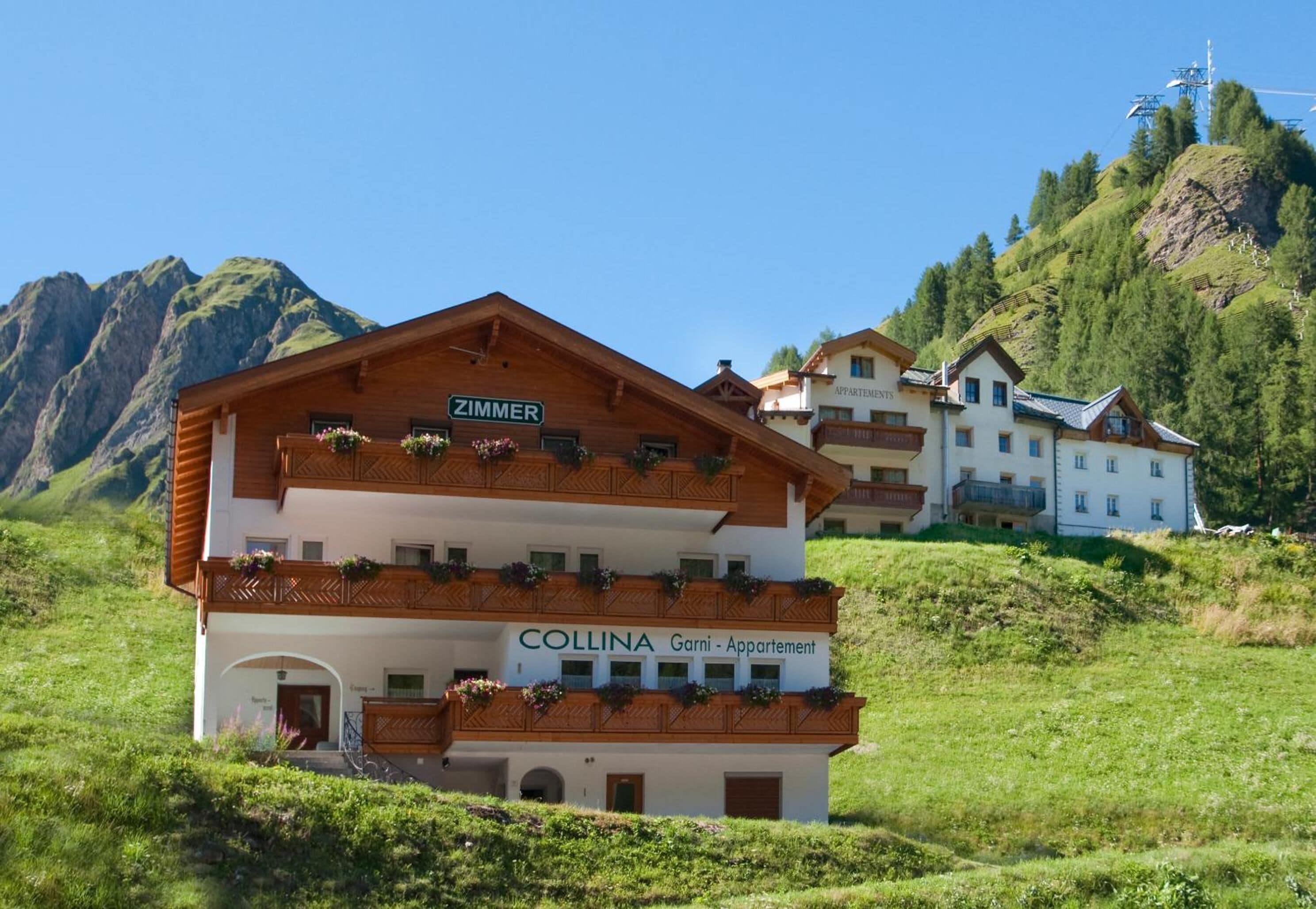 A multi-story building with balconies, labeled "Zimmer" and "Collina Garni - Appartement," set against a mountainous backdrop.