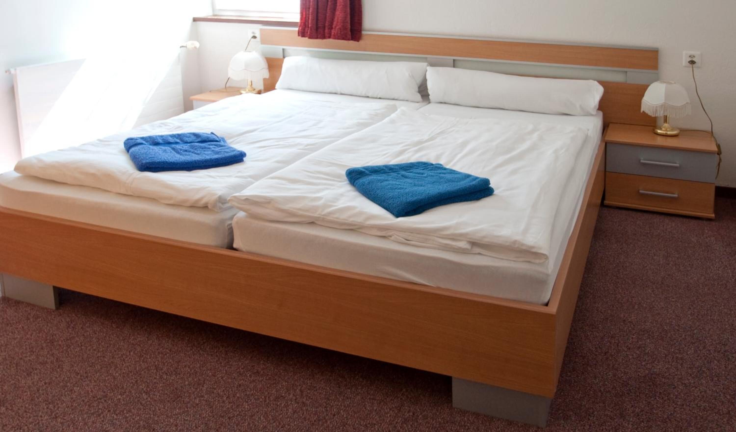 A double bed with white linens and blue towels, a wooden headboard, bedside tables with lamps, and a carpeted floor.
