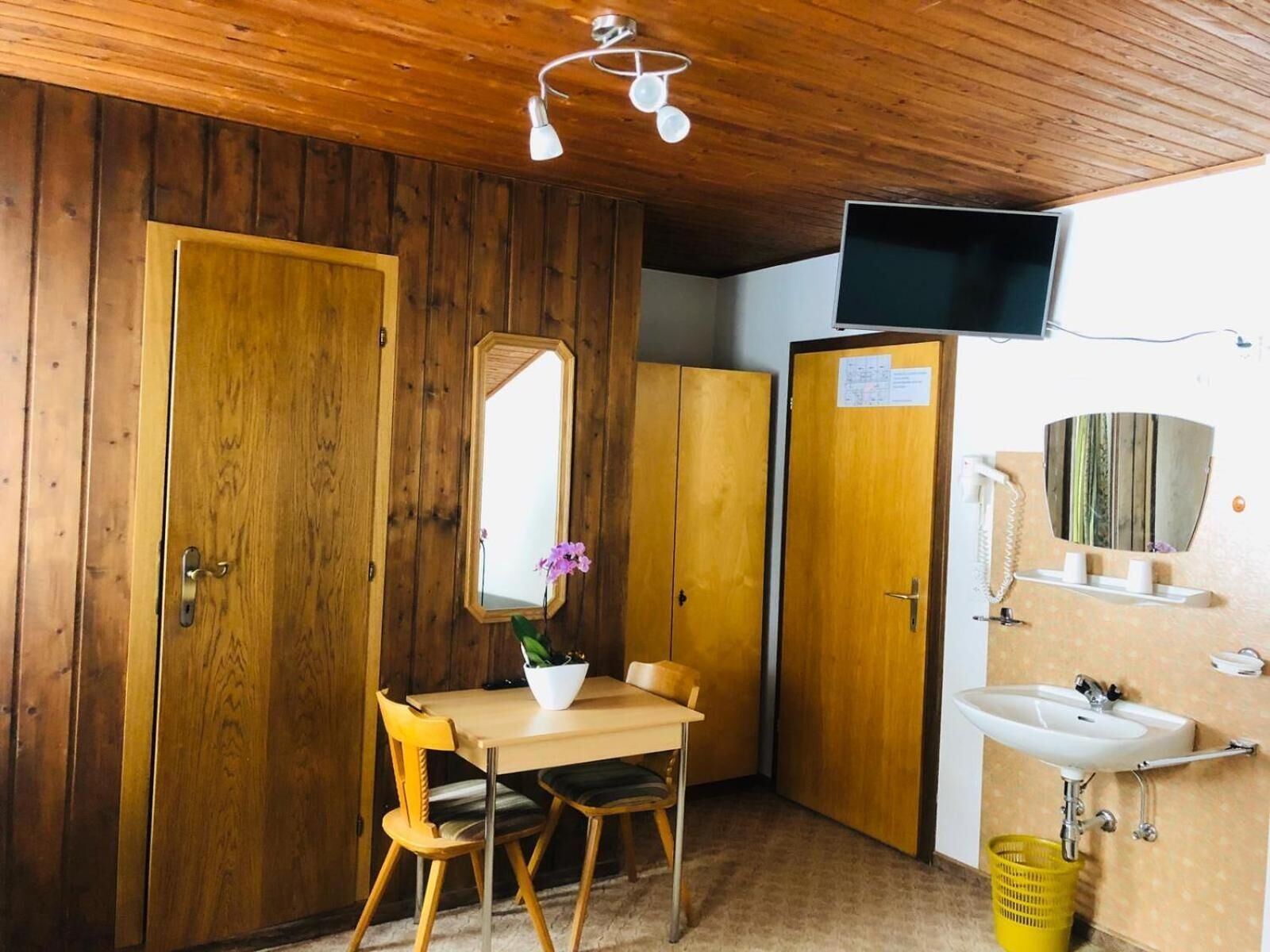 Une petite pièce aux dimensions réduites, avec des murs et un plafond en bois, comprenant un bureau en bois avec une chaise, un miroir, un téléviseur fixé au mur et un lavabo avec un panier jaune.