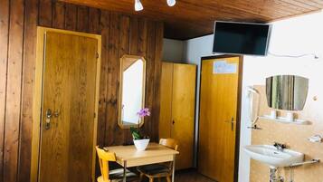 Une petite pièce aux dimensions réduites, avec des murs et un plafond en bois, comprenant un bureau en bois avec une chaise, un miroir, un téléviseur fixé au mur et un lavabo avec un panier jaune.