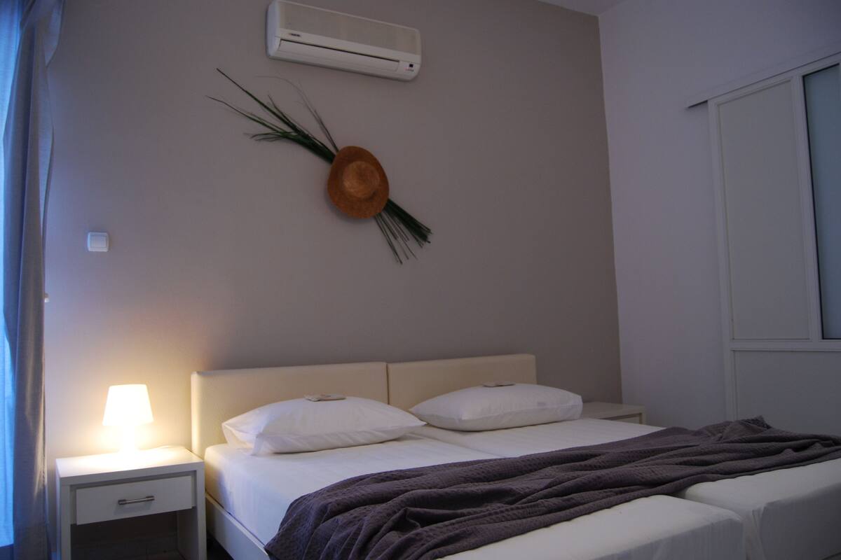 Hotelkamer met een bed, nachtkastje met lamp, wandairconditioner en een decoratieve plant.