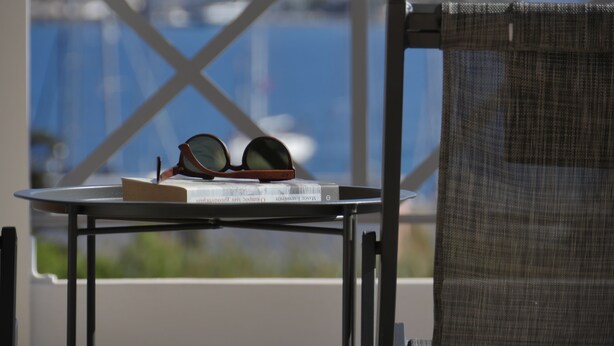 A table with a pair of sunglasses and a book, a chair with a mesh back, and a view of a marina through a window with a white cross pattern. - Galini Rooms & Apartments Syros (Syros)