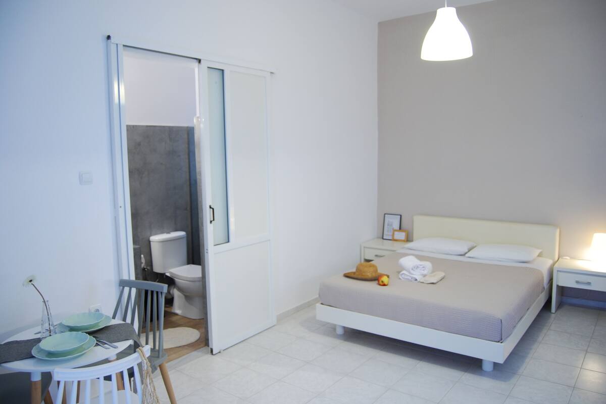 Een slaapkamer met een bed, nachtkastje en een stoel. De aangrenzende badkamer heeft een toilet en een wastafel.