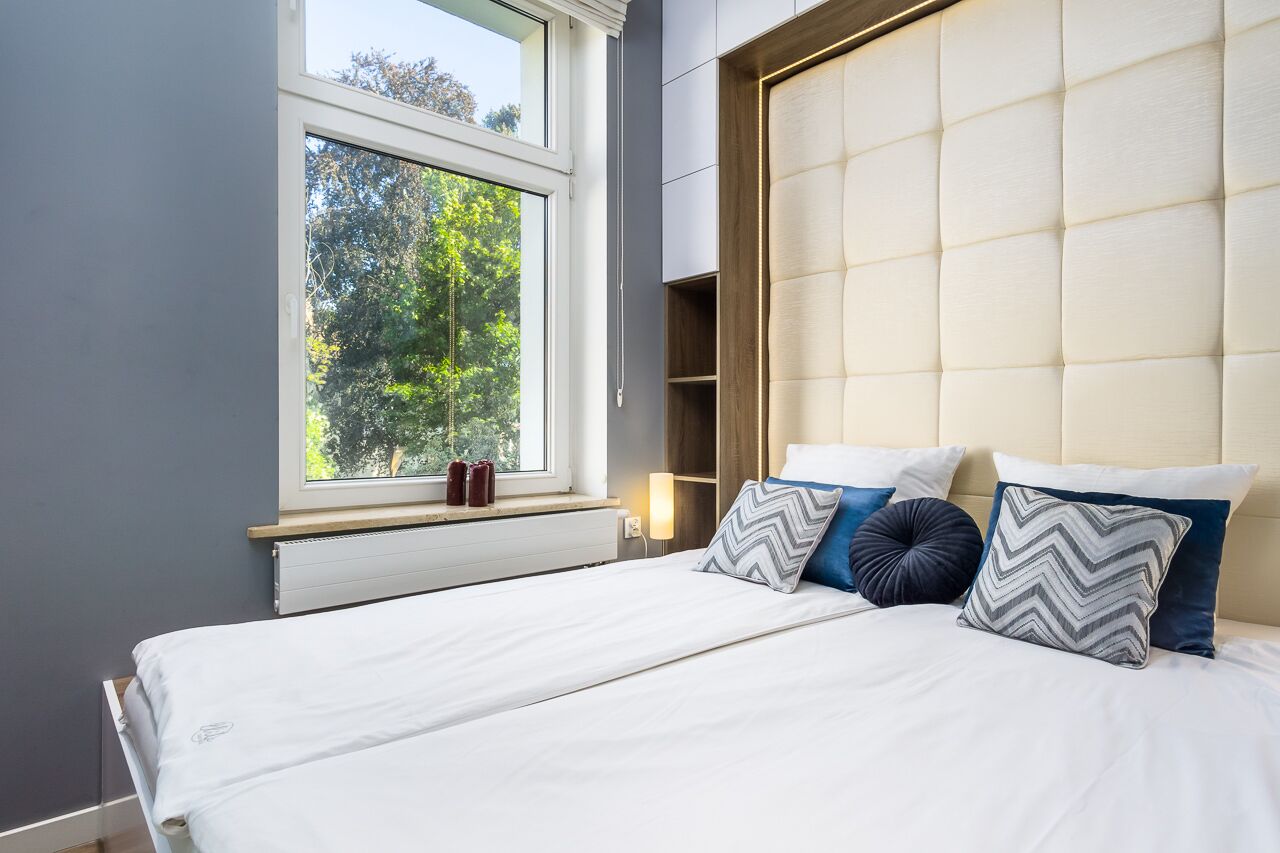Ein modernes Schlafzimmer mit einem großen Fenster, einem Bett mit weißer Bettwäsche und einem gepolsterten Kopfteil.