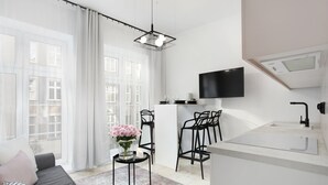 A modern living room with a sofa, a coffee table with flowers, a kitchen area with a TV, and a large window with curtains.