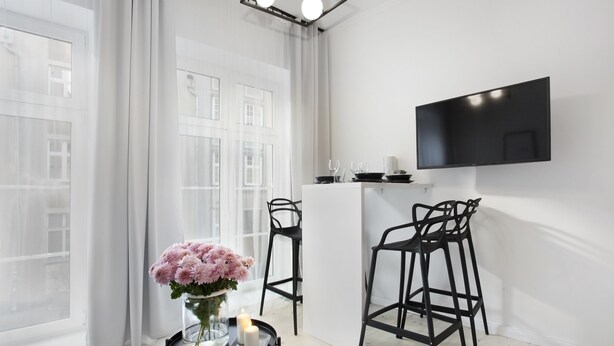 A modern room with a large window, a flat-screen TV mounted on the wall, two black bar stools, a small round table with a vase of flowers, and a candle.