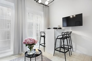 Une chambre moderne dotée d’une grande fenêtre, d’un téléviseur à écran plat fixé au mur, de deux tabourets de bar noirs, d’une petite table ronde ornée d’un vase de fleurs et d’une bougie.
