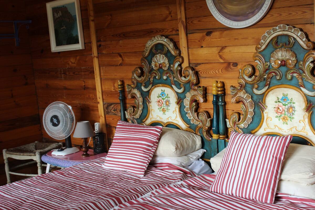 Puuinen mökkimäinen huone, jossa on sänky, tuoli, tuuletin ja lamppuvarustettu pöytä.