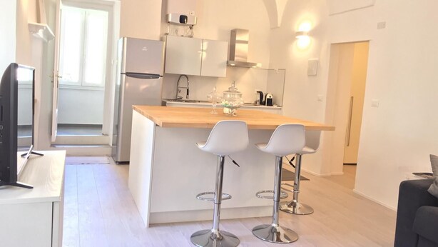 A modern kitchen with a central island, white cabinetry, and stainless steel appliances. There are two bar stools at the island and a television on the left. - Happy Holiday Sanremo (Sanremo)