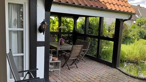 Een overdekt terras met een geplaveid vloertje, een tafel met stoelen en uitzicht op groen.