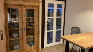 Een houten kast met glazen deuren, gevuld met servies, een witte deur met glaspanelen, en een houten tafel met metalen poten.