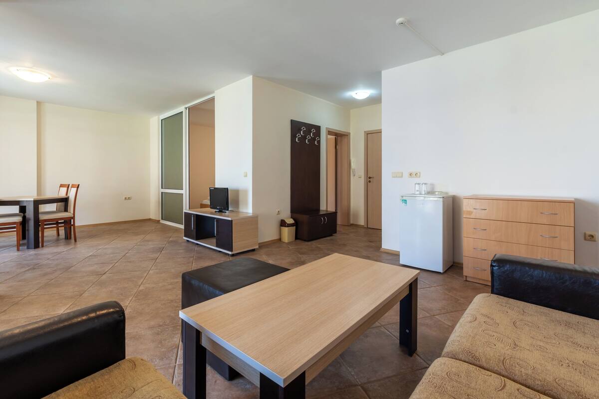 A modern living room with a wooden table, black and beige sofas, a TV, and a kitchen area with cabinets and a refrigerator.