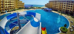 A large swimming pool with a water slide, surrounded by hotel buildings and a clear view of the ocean. - Midia Family Resort (Pomorie)