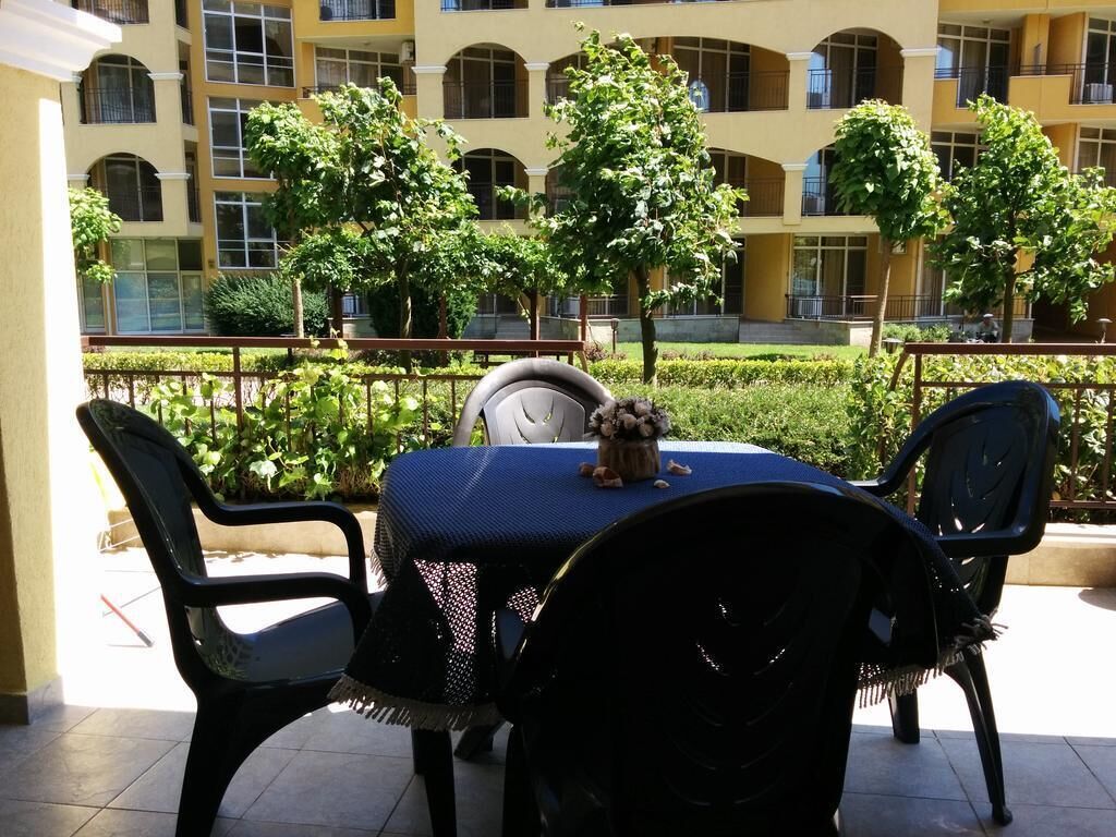Eine Terrasse mit einem runden Tisch und zwei Stühlen, umgeben von Grünpflanzen und einem Gebäude mit zahlreichen Fenstern im Hintergrund.