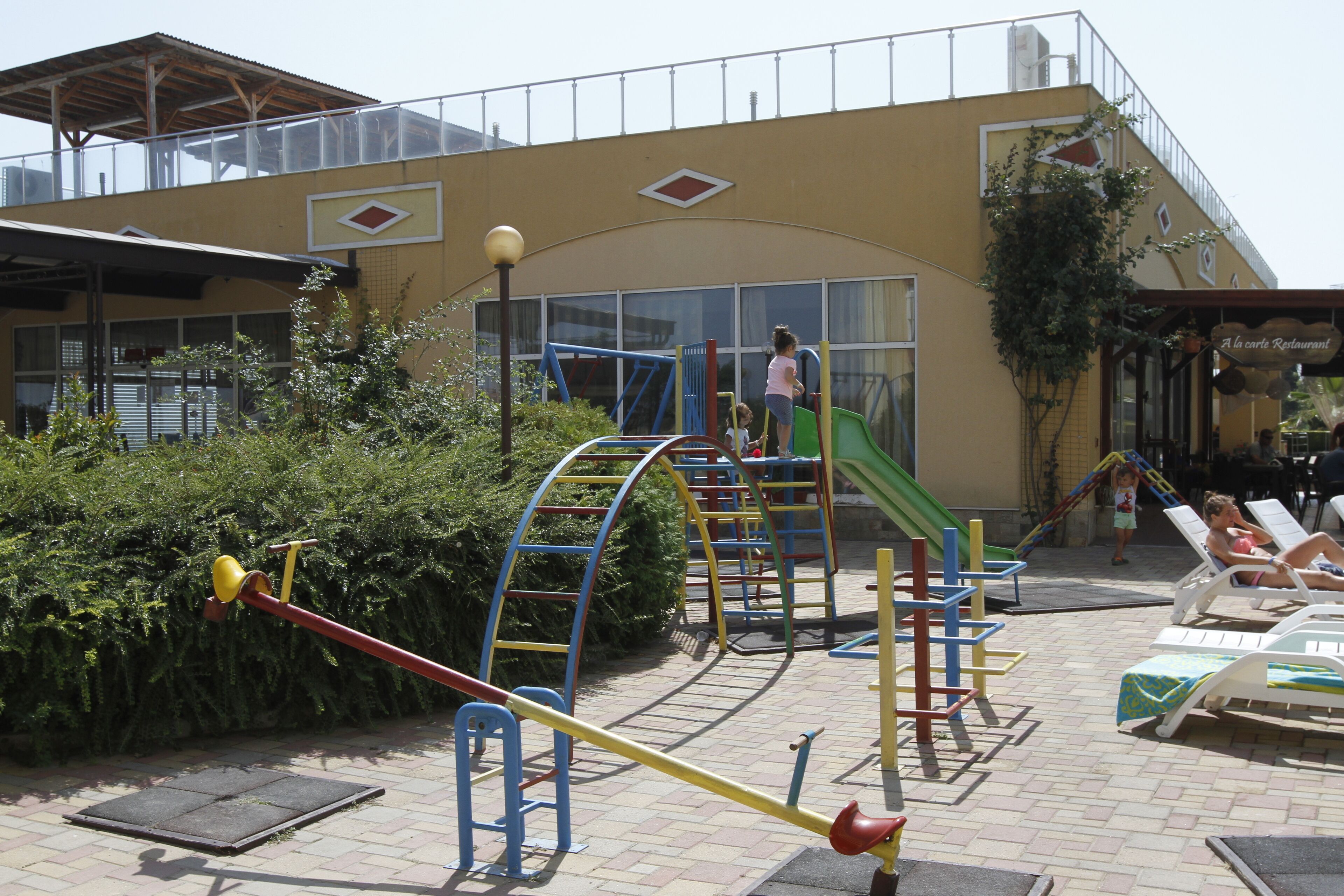 A playground with various play structures, a slide, and a building with a restaurant sign in the background.