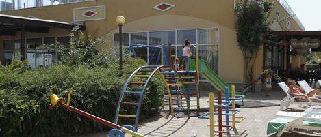 A playground with various play structures, a slide, and a building with a restaurant sign in the background.