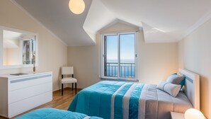 Una camera da letto con un'ampia finestra, un letto con biancheria azzurra e grigia, un comò bianco e una sedia.