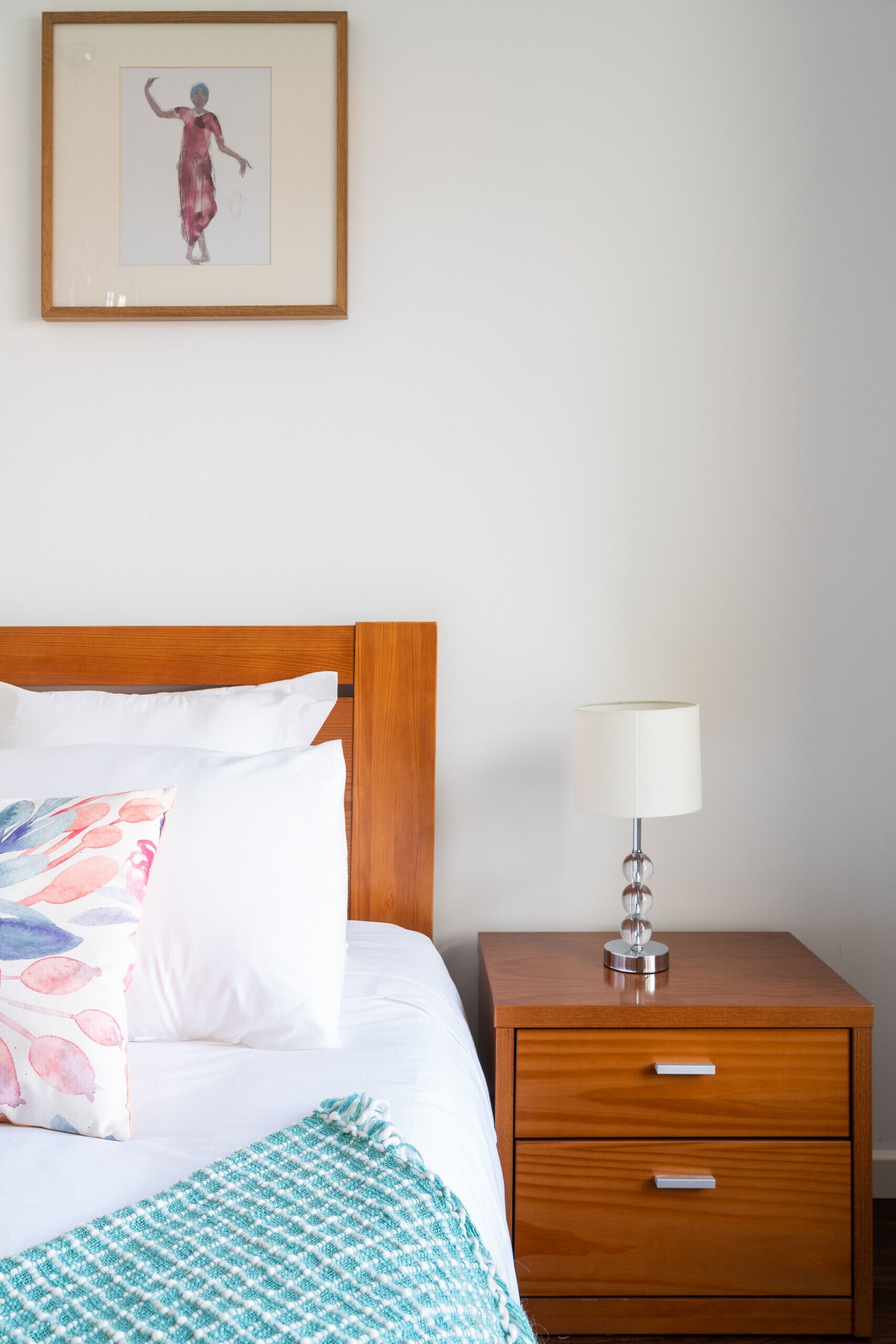 Een net opgemaakt bed met witte en kleurrijke kussens, een houten hoofdbord, een bedlamp en een klein houten nachtkastje met lades.