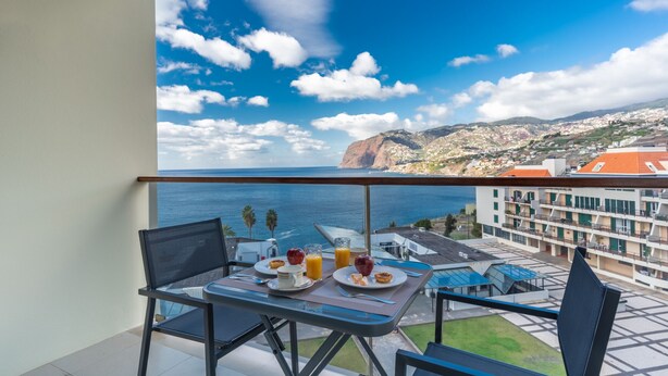 Varanda com mesa posta para o café da manhã, com vista para um centro urbano costeiro com edifícios e um penhasco.