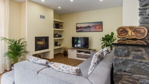 A living room with a grey sofa, a television, a fireplace, and a bookshelf with wine bottles. - Villa Teresa/1 by Atlantic Holiday (São Vicente)