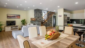 A modern living room with a stone fireplace, a dining table with a fruit basket, and a kitchen area with a refrigerator and stove. - Villa Teresa/1 by Atlantic Holiday (São Vicente)