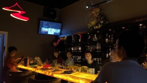 Een bar met een lange toonbank, een televisie aan de muur en planken met flessen en glazen.