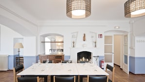 A dining area with a long table set for six, flanked by two chandeliers, a fireplace, and a kitchen in the background. - Santuari de Terrades (Terrades)