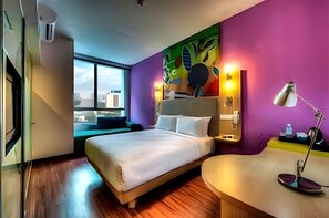 Une chambre d’hôtel avec un lit, un bureau, une lampe et une fenêtre donnant sur des immeubles.