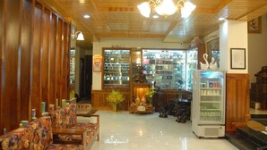A lobby with wooden paneling, a chandelier, a seating area with floral-patterned sofas, a glass display case with items, and a vending machine. - 7S Hotel Le Na (Cam Ly - Da Lat)