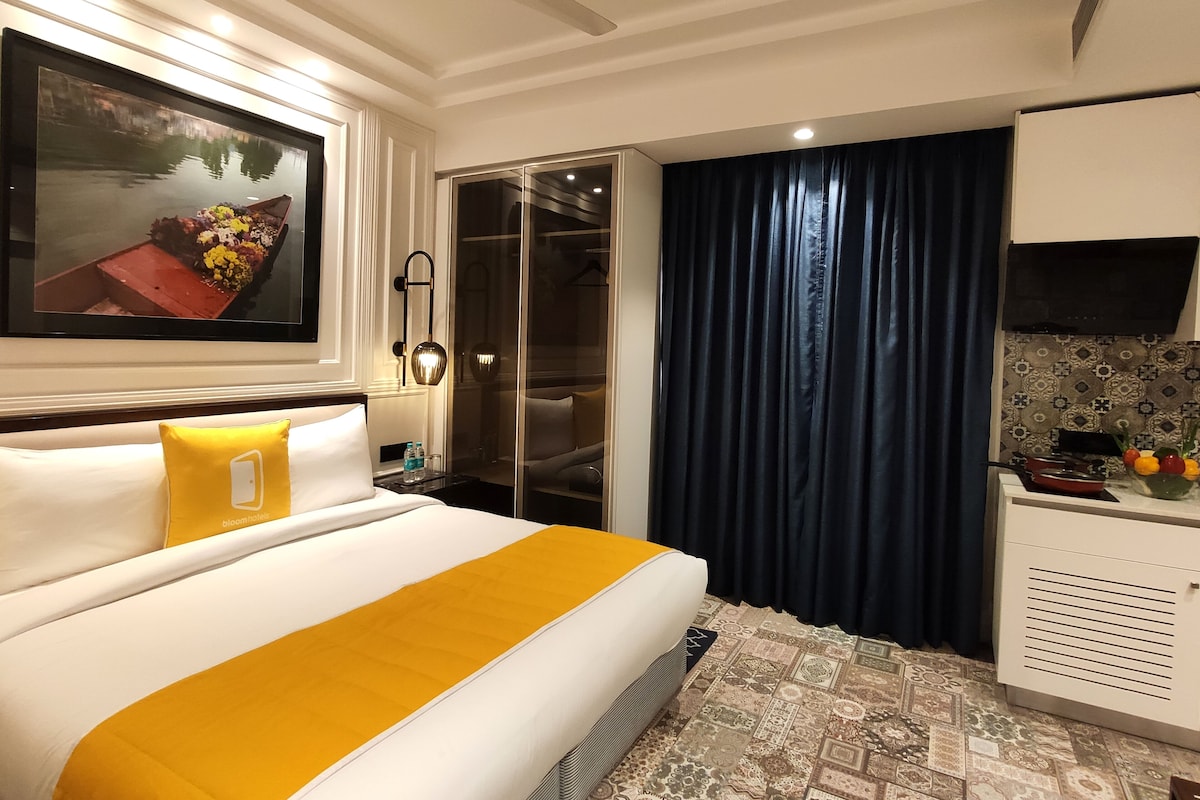 A hotel room with a bed, a nightstand, a mirror, a wardrobe, a window with curtains, and a kitchenette area.