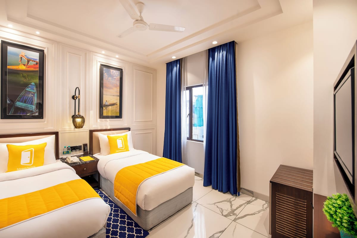 A hotel room with two beds, a ceiling fan, artwork on the walls, a window with blue curtains, and a small plant.