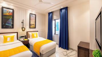 A hotel room with two beds, a ceiling fan, artwork on the walls, a window with blue curtains, and a small plant.