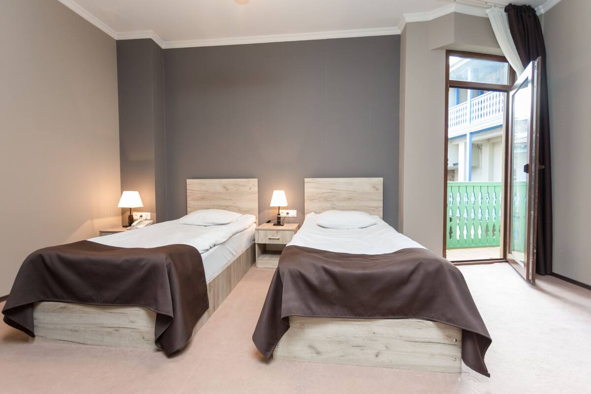 A hotel room with two beds, bedside lamps, a wooden headboard, a balcony with a green fence, and a sliding glass door.