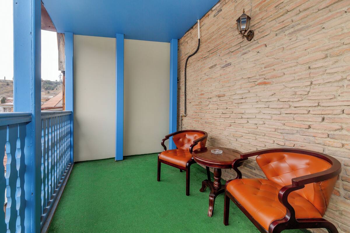 A balcony with artificial green flooring, two wooden chairs, a small round table, and a brick wall with a hanging light fixture.