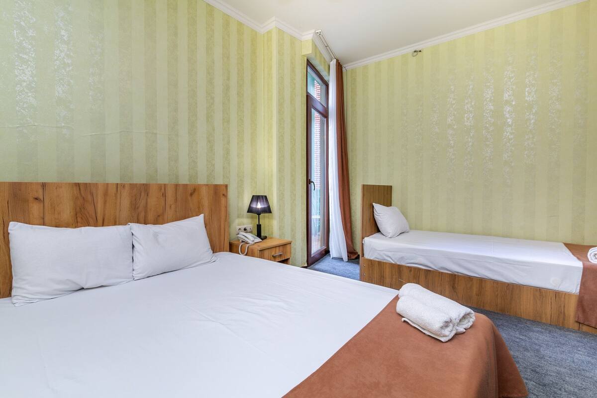 A hotel room with two beds, a wooden headboard, a nightstand with a lamp, and a window with curtains.
