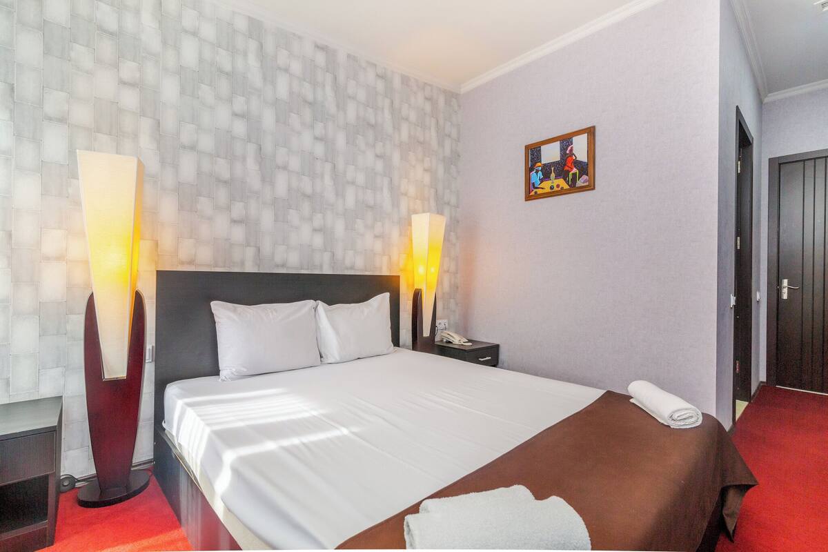 A hotel room with a large bed, two bedside lamps, a nightstand, a painting on the wall, and a door to the right.
