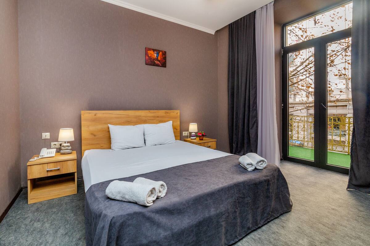 A hotel room with a large bed, a nightstand with a lamp, a telephone, and a small table with a flower.