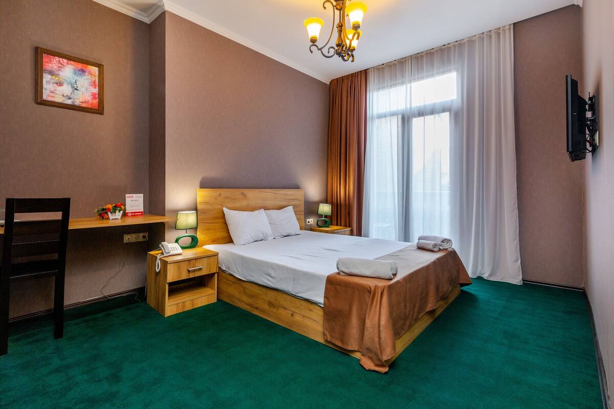 A hotel room with a wooden bed, green carpet, a desk with a chair, a TV on the wall, and a window with curtains.