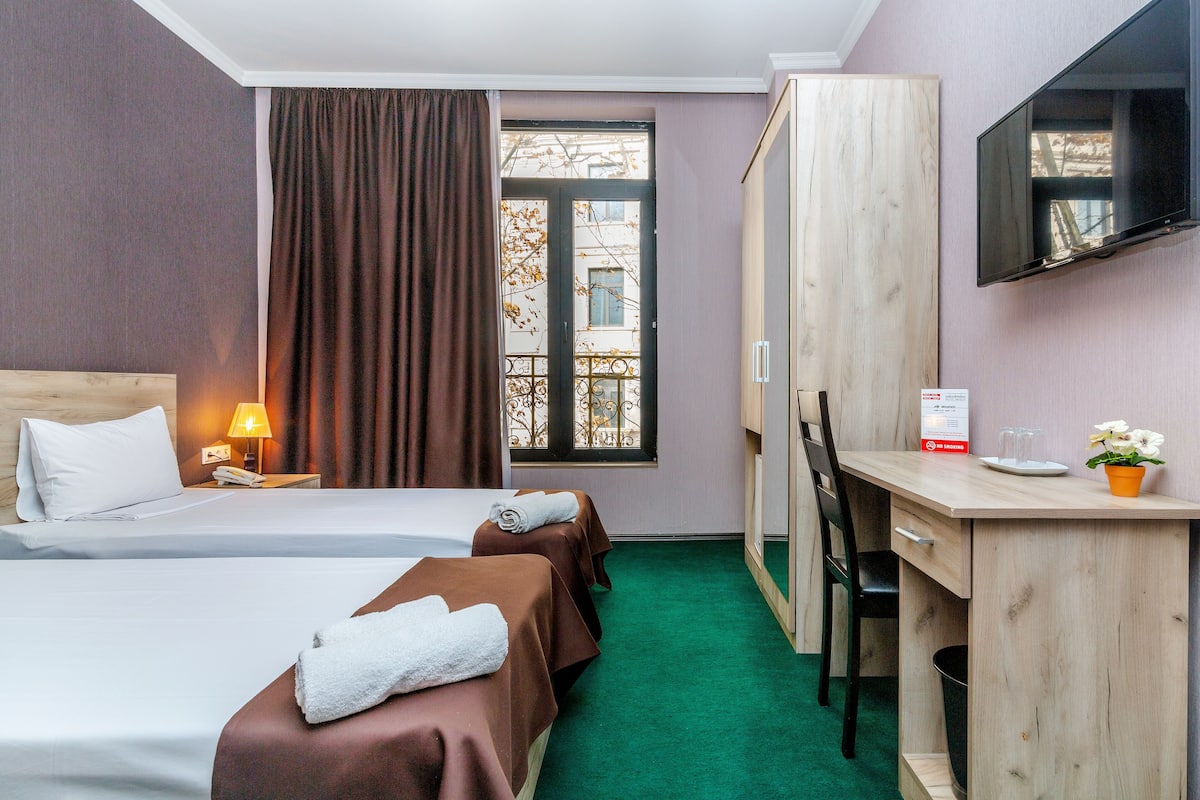 A hotel room with two beds, a desk, a TV, and a window with a view of buildings.