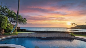 Una piscina con vistas al océano y al atardecer, rodeada de vegetación y palmeras.