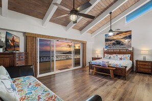 Un dormitorio con cama, banca, mesita de noche, ventilador de techo de madera y una ventana grande con vistas a la playa.